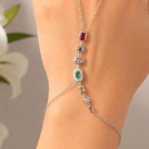 May include: A silver hand chain bracelet featuring a delicate chain that connects a wrist band to a finger loop. The central chain is adorned with a series of multi-coloured gemstones in various shapes, including red, blue, pink, green, and yellow.