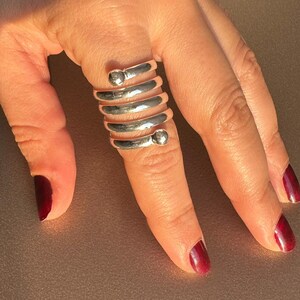 Könnte beinhalten: Ein silberfarbener Ring umschlingt einen Finger und weist ein spiralförmiges Design mit kleinen, abgerundeten Akzenten auf. Der metallische Glanz des Rings kontrastiert mit dem Hautton und dem dunkelroten Nagellack.