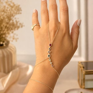 Könnte beinhalten: Eine goldfarbene Handkette mit einer feinen Kette, die einen Ring mit einem Armband verbindet. Die mittlere Kette ist mit einer Reihe kleiner, mehrfarbiger Edelsteine in verschiedenen Formen verziert, darunter rote, blaue, violette, grüne und rosa.