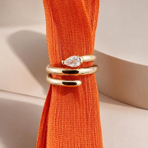 Puede incluir: Un anillo abierto de tono dorado con una banda de metal lisa y pulida que se envuelve alrededor del dedo. Un extremo del anillo está acentuado con una gema transparente en forma de pera, mientras que el otro termina en una punta redondeada.