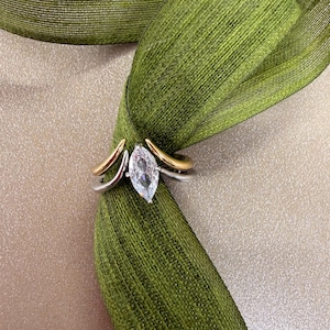 May include: A two-tone ring featuring a marquise-cut clear gemstone set in the centre. The band is constructed from a silver-coloured metal and a gold-coloured metal, which curve upwards to meet the central stone in a bypass-style design.