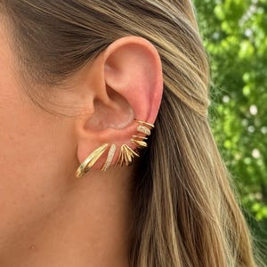 May include: A collection of gold-toned ear jewelry consisting of multiple pieces. The set includes a smooth curved earring, a pavé-set crystal hoop, a fan-shaped earring, and several stacked ear cuffs, some featuring small clear crystals.