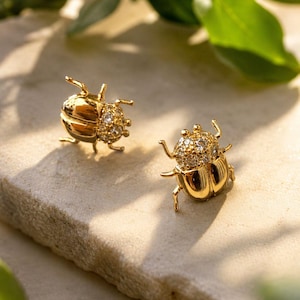 Könnte beinhalten: Ein Paar goldfarbene Ohrstecker in Käferform. Die Ohrringe haben ein detailliertes Design mit kleinen, funkelnden, klaren Steinen auf Kopf und Körper. Sie werden auf einer hellen, strukturierten Oberfläche mit grünen Blättern im Hintergrund präsentiert.