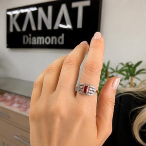 May include: A rectangular ruby gemstone ring with a halo of small diamonds. The ring is set in a silver band. The ring is on a person's finger. The background includes the text "KANAT Diamond".