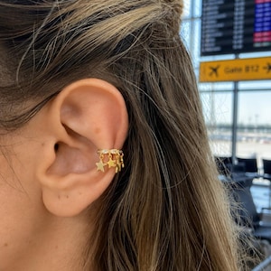 May include: Gold ear cuff featuring a row of small clear stones and dangling gold stars. The ear cuff is worn on the upper ear. The background is blurred, but appears to be an airport terminal.