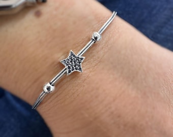 May include: A silver bracelet featuring a star charm with black embellishments. The bracelet has a delicate chain design with small, round beads. A silver watch is visible in the background.