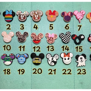 May include: A collection of 24 colorful shoe charms featuring various designs. The charms include Mickey Mouse, Minnie Mouse, and other character-themed designs. The charms are numbered 1 through 24 and are arranged on a light green surface.