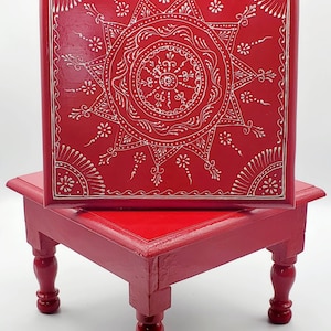 May include: A red wooden stool with four turned legs and a square top. The top of the stool is decorated with a white mandala design.