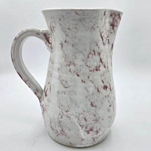 May include: White ceramic pitcher with a handle and a wide spout. The pitcher has a speckled pattern of maroon and white. The handle is curved and attached to the side of the pitcher. The pitcher is likely used for serving beverages.
