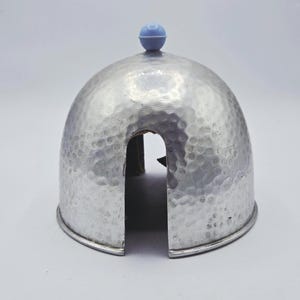 May include: A dome-shaped silver-colored metal food cover with a hammered texture. The cover features a single arched opening at the base and a small, round, light blue knob handle centered at the very top of the dome structure.