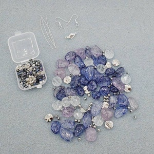 May include: A close-up of a jewelry making kit with a variety of purple and clear glass beads, silver beads, and earring findings. The beads are in a small plastic container and the findings are scattered on a gray surface.
