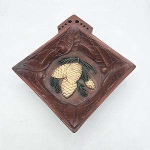 May include: A diamond-shaped brown wall plaque featuring a central circular relief of three pinecones in gold and green. The border is decorated with carved leaf patterns, and the top section includes a small tab with six drilled holes.