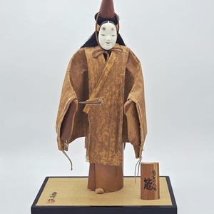 May include: A Japanese Noh theater doll featuring a white mask, black hair, and a tall brown pointed hat. The doll wears a textured brown robe with a mottled pattern and holds a small fan. It stands on a rectangular wooden base with a small sign.