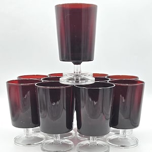 May include: A set of ten matching drinking glasses featuring deep red, translucent bowls and clear glass stems with circular bases. The glasses are arranged in a tiered formation, with one glass elevated on top of the others, showcasing a uniform design.