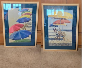 May include: Two framed beach scene prints. One print shows colorful beach umbrellas and chairs on a sandy beach. The other print shows beach umbrellas and chairs on a sandy beach. Both prints have a blue mat and a light wood frame.