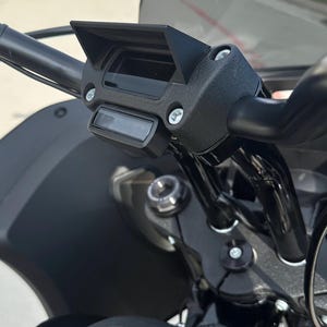 May include: Close-up of a black motorcycle handlebar with a black visor and a digital display. The motorcycle has a black fuel tank with the text "FXLR ST" printed on it.
