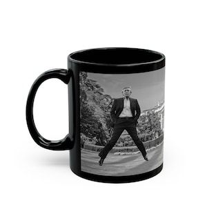 May include: Black coffee mug with a white image of a person in a suit standing in front of a building. The person is making a jumping pose with their legs spread wide.