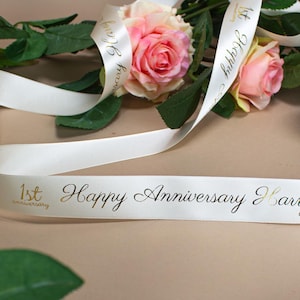 May include: White satin ribbon with gold lettering that reads "1st anniversary Happy Anniversary" and pink roses.