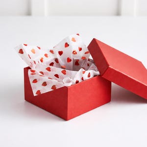 May include: A red gift box with the lid open, showing white tissue paper decorated with red hearts. The box is square and has a matte finish. The tissue paper is crumpled and spilling out of the box.