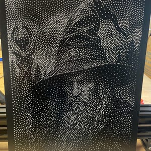 以下が含まれることがあります： 多数の小さな反射する銀色のスタッドで構成された点描画スタイルの長方形の黒いパネル。高い帽子をかぶり、杖を持った髭を生やした人物が、高いコントラストで描かれています。