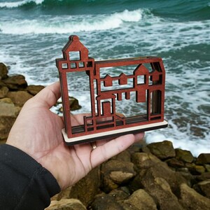 May include: A wooden cutout of a lighthouse and houses, painted in shades of brown and red. The cutout is held in a hand against a backdrop of ocean waves.
