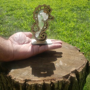 May include: A wooden seahorse figurine with a cutout design. The seahorse is filled with small pieces of sea glass in shades of green and white. The figurine is standing on a small wooden base.