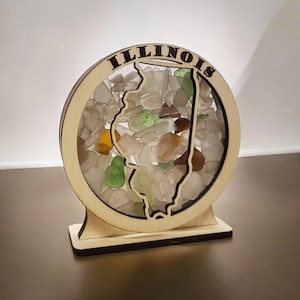May include: A wooden cutout of the state of Illinois filled with colorful sea glass. The cutout is mounted on a wooden base and the words "ILLINOIS" are engraved around the top of the cutout.