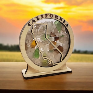 May include: A wooden cutout of the state of California filled with sea glass. The cutout is mounted on a wooden base and has the word "CALIFORNIA" written around the edge.