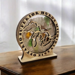 May include: Wooden Great Lakes map with a circular frame and a clear center filled with colorful sea glass. The names of the Great Lakes are engraved around the frame: Superior, Huron, Michigan, Erie, Ontario.