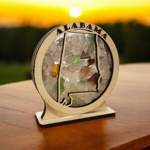 May include: A wooden cutout of the state of Alabama filled with colorful sea glass. The cutout is mounted on a wooden base and has the word "ALABAMA" written around the top.