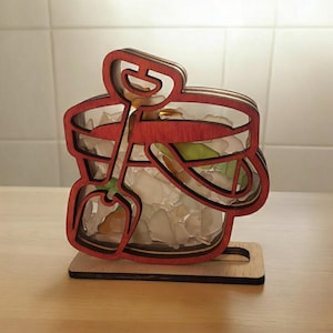 May include: A wooden cutout of a bucket and shovel filled with colorful glass beads. The bucket is red and the shovel is brown. The base is made of wood.