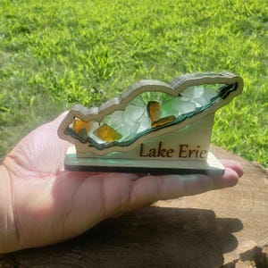 May include: Wooden Lake Erie outline filled with clear and amber glass pieces. The base is wood with the words "Lake Erie" engraved in brown.