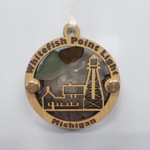 May include: A wooden ornament featuring a lighthouse and buildings, with the text "Whitefish Point Light Michigan" engraved around the edge. The center is filled with sea glass. Includes a string for hanging.