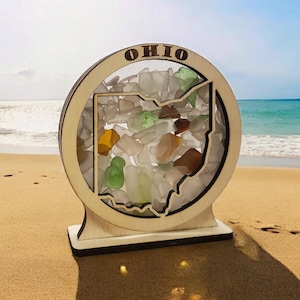 May include: A wooden cutout of the state of Ohio filled with sea glass. The cutout is mounted on a round wooden base with the word "OHIO" engraved on the top. The base is sitting on a sandy beach.
