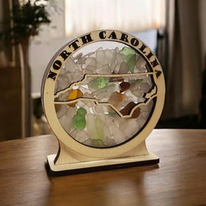 May include: A wooden North Carolina state-shaped display filled with sea glass. The state outline is cut out within a circular frame with "NORTH CAROLINA" engraved around the edge. The sea glass is in shades of white, green, and brown.