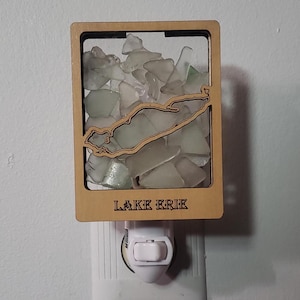 May include: A wooden night light with a clear glass insert filled with small pieces of sea glass. The glass is shaped to resemble the outline of an island. The night light is plugged into an electrical outlet and has a switch to turn it on and off. The text "LAKE ERIE" is printed on the bottom of the night light.