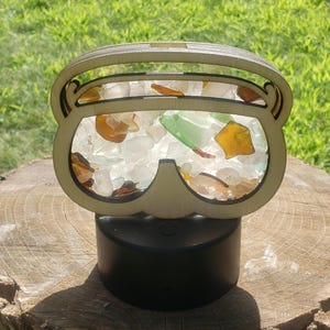 May include: A wooden cutout of a pair of sunglasses with a light wood finish. The sunglasses are filled with colorful sea glass, including clear, green, and brown pieces.