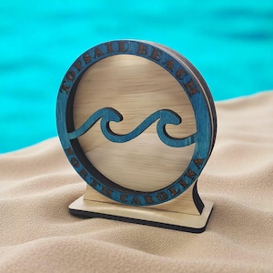 May include: Wooden tabletop decoration featuring a blue-painted circular frame with the words "TOPSAIL BEACH NORTH CAROLINA". The center has a wave design. The decoration is on a wooden base, set against a sandy background.