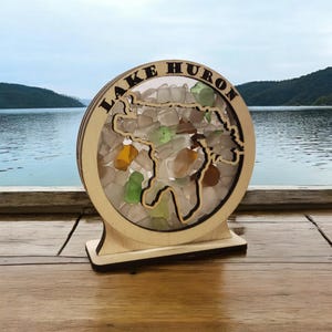 May include: A wooden cutout of Lake Huron filled with colorful sea glass. The cutout is mounted on a wooden base and reads "LAKE HURON".
