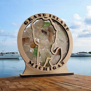 May include: A wooden cutout of Grand Island, Michigan, filled with sea glass. The cutout is on a wooden base and the words "Grand Island Michigan" are engraved on the base.