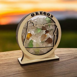 May include: A wooden cutout of the state of Oregon filled with colorful sea glass. The cutout is mounted on a wooden base and has the word "OREGON" written around the outside.