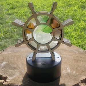 May include: A wooden ship's wheel with a light inside, filled with sea glass. The wheel is on a black base with a power switch.