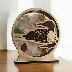 May include: A wooden circular ornament with a loon silhouette and a clear resin insert filled with colorful glass pieces. The ornament is on a wooden stand.