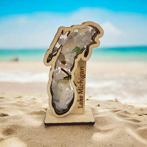 May include: A wooden Lake Michigan art piece filled with sea glass. The wooden frame is in the shape of the lake, with the words "Lake Michigan" engraved on the side. The background is a beach with blue water and sky.