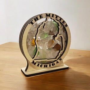 May include: A wooden cutout of the state of Michigan filled with colorful sea glass. The cutout is surrounded by a circle with the words "The Mitten Michigan".