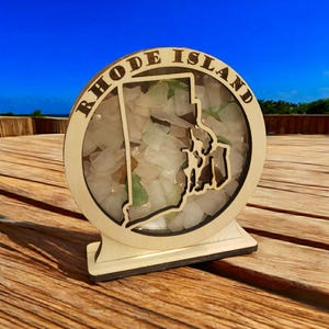 May include: A wooden cutout of the state of Rhode Island filled with small pieces of sea glass. The cutout is mounted on a wooden base and has the words "Rhode Island" inscribed around the edge.
