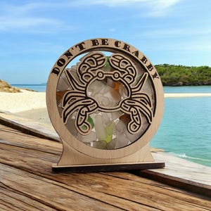 May include: A wooden sign with a round cutout featuring a crab design and the text "DON'T BE CRABBY". The cutout is filled with small pieces of sea glass.