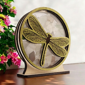 May include: A wooden decorative stand with a round, clear glass insert. The stand features a laser-cut dragonfly design with a yellow and brown color scheme. The glass insert is filled with small, white, and clear stones.
