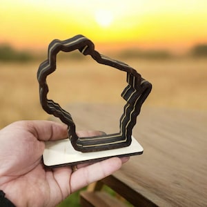 May include: A wooden cutout of the state of Wisconsin, layered and standing on a small wooden base. The cutout is dark brown with thin gold lines.