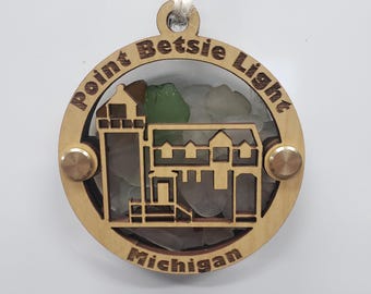 Puede incluir: Adorno de madera circular con un diseño de faro y edificio, con el texto "Point Betsie Light Michigan". El adorno tiene un centro transparente lleno de vidrio de mar, detalles en latón y una cuerda para colgar.